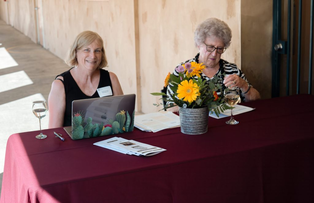 Two people seated at a check-in table with a laptop and a vase of flowers.