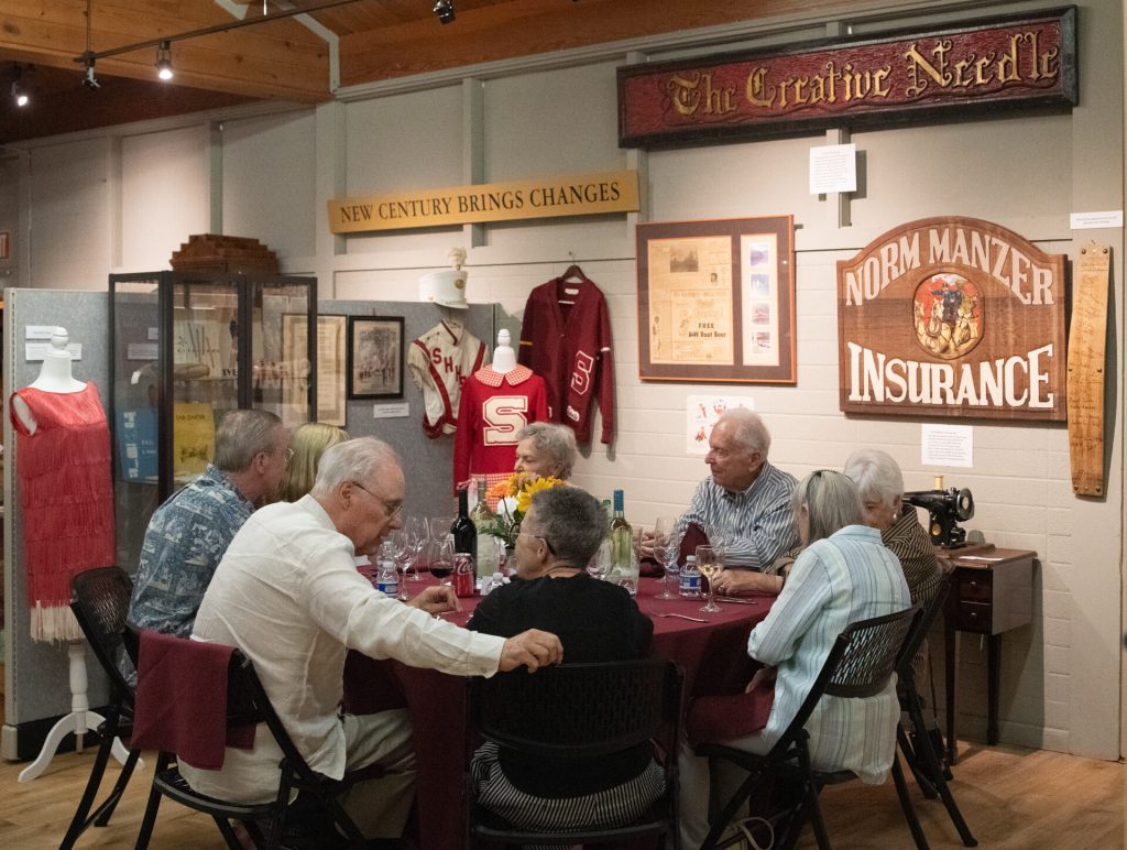 Guests dine at round tables beneath museum signs reading The Creative Needle and Norm Manzer Insurance.